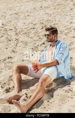 Gut aussehender bärtiger Mann in Sonnenbrille sitzt am Sandstrand mit einem Glas Alkohol cocktail Stockfoto