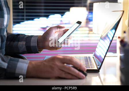 Mann mit Smartphone und Laptop spät in der Nacht. Unternehmer oder Kaufmann in der modernen Arbeit im Büro. Die Lichter der Stadt im Hintergrund. Stockfoto