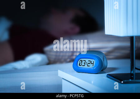 Schließen Sie bis zu digitaler Wecker auf dem Nachttisch im Schlafzimmer. Mann schlafen im Bett in den Hintergrund. Verträumt blur Effekt. Stockfoto