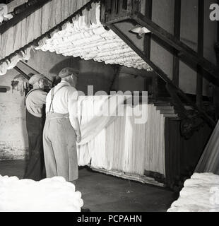 1950, historische Mühle, männliche Arbeitnehmer in Overalls und tragen Kappen, innen prüfen Rollen aus Gewebe aus Leinen, Nordirland. Stockfoto