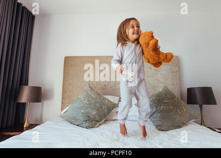 Fröhliches kleines Mädchen springen auf Bett mit Teddybär. Kind, Mädchen spielen auf dem Bett in Ihrem Schlafzimmer und lächelnd. Stockfoto
