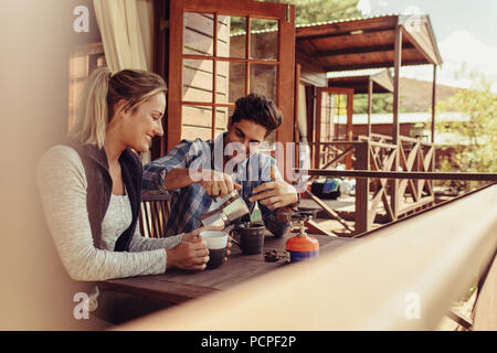 Paar außerhalb einer Ferienhütte und Kaffee sitzen. Mann Kaffee gießen in Schale mit seiner Freundin durch Sitzen. Stockfoto