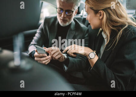 Junge Frau mit älteren Geschäftsmann mit Mobiltelefon, während sie von einem Auto unterwegs. Geschäftsleute können Sie über Smart Phone im Taxi. Stockfoto