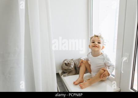 Süß lächelnd Kind mit grauen Britisch Kurzhaar Katze auf Fensterbank zu Hause Stockfoto