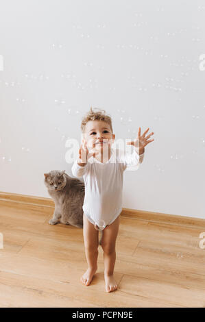 Lächelnd Kleinkind Junge in Weiße Strampler fangen Seifenblasen mit graue Katze in der Nähe von zu Hause. Stockfoto