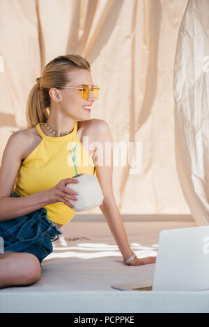 Schönen lächelnden jungen Frau in Sonnenbrille holding Sommer coconut Cocktail und Wegsehen Stockfoto