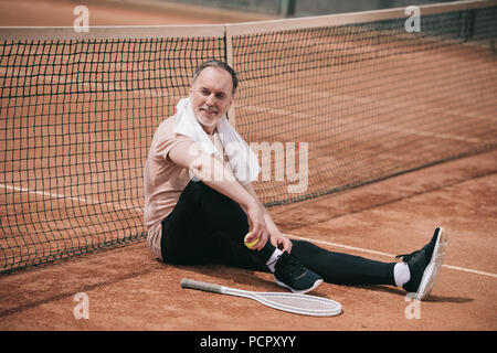 Lächelnd älterer Mann mit Handtuch- und Tennisausrüstung ruht in der Nähe von Netz auf dem Tennisplatz Stockfoto