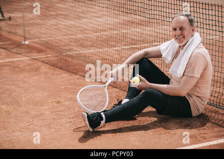 Lächelnd älterer Mann mit Handtuch- und Tennisausrüstung ruht in der Nähe von Netz auf dem Tennisplatz Stockfoto