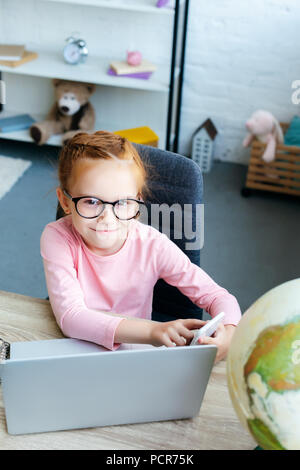 Hohe Betrachtungswinkel und niedlichen Rothaarigen Schulkind in Brillen in die Kamera lächeln, während Sie ihr Smartphone und Laptop Stockfoto