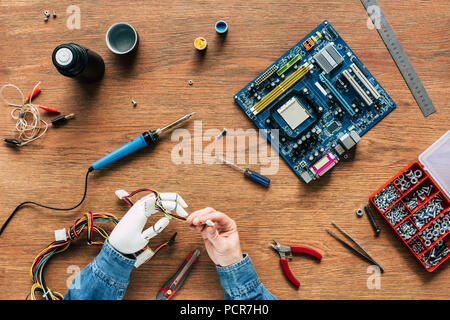 Zugeschnittenes Bild des Menschen mit roboterhand Kabel Holding mit Werkzeugen an den hölzernen Tisch Stockfoto