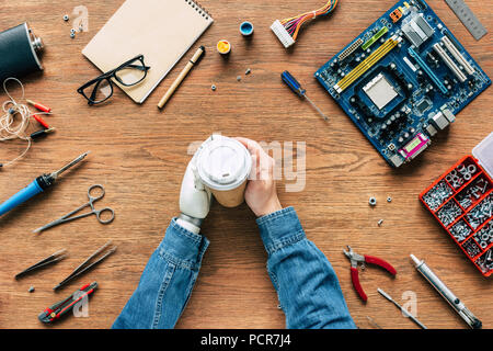 Zugeschnittenes Bild elektronischer Ingenieur mit prothetischen Arm holding Pappbecher auf Tabelle mit Tools Stockfoto