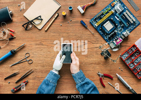 Zugeschnittenes Bild elektronischer Ingenieur mit prothetischen Querlenker mit Smartphone bei Tisch umgeben von Tools Stockfoto