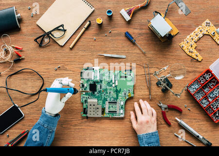 Zugeschnittenes Bild elektronischer Ingenieur mit roboterhand zur Festsetzung der Hauptplatine durch Lötkolben am Tisch umgeben von Tools Stockfoto