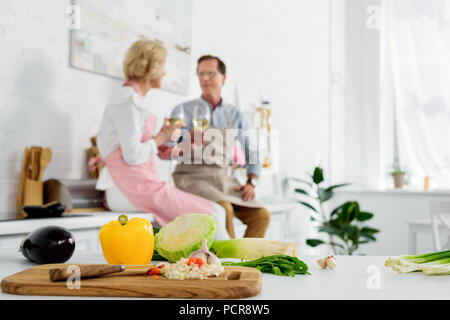 Nahaufnahme von frischem Gemüse in Holz Schneidebrett und Senior paar Wein trinken hinter in der Küche Stockfoto