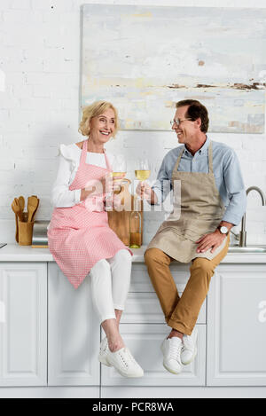 Gerne älteres Paar in Schürzen holding Gläser Wein, während sitzen gemeinsam in der Küche Stockfoto