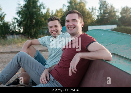 Zwei jungen kaukasischen Männern sitzen auf Sandstrand und lächelnd. Die besten Freunde von der Hochschule Konzept. Stockfoto