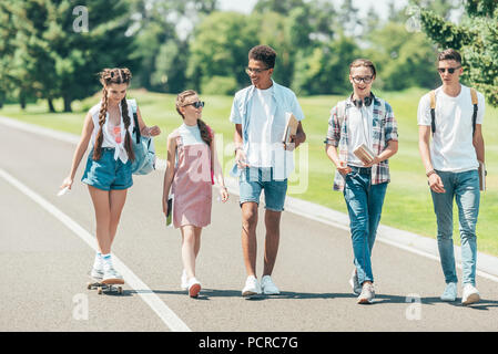 Multiethnische gruppe Teenager mit Bücher- und Skateboard zu reden und gemeinsam zu Fuß in Park Stockfoto