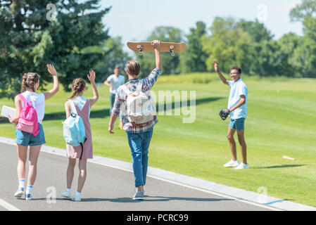 Jugendliche gehen mit Bücher- und Skateboard in Park- und Gruß Klassenkameraden spielen Baseball Stockfoto