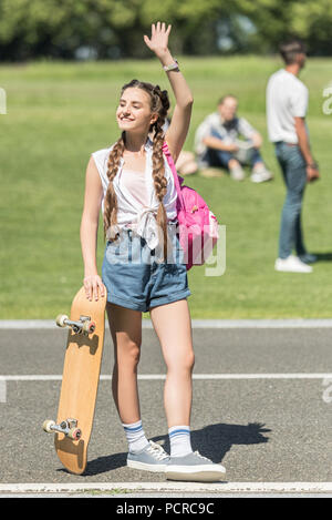 Smiling teenage Mädchen mit Skateboard und Rucksack Hand winken und weg schauen in Park Stockfoto