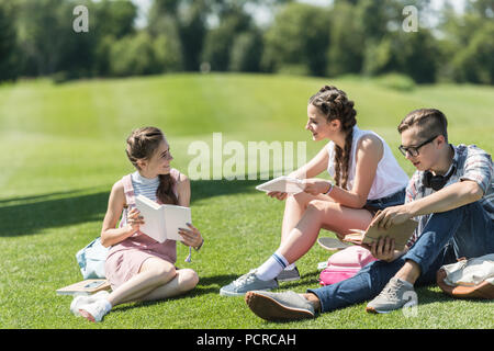 Smiling teenage Studenten mit Büchern und digitalen Tablet sitzen auf Gras und gemeinsam studieren in Park Stockfoto