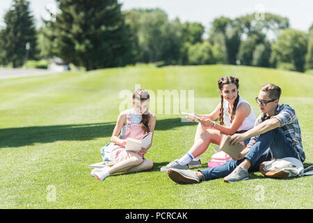 Smiling teenage Studenten sitzen auf Gras und Studieren mit Büchern und digitalen Tablet in Park Stockfoto