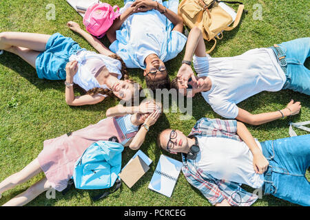 Blick von oben auf die multiethnische halbwüchsigen Klassenkameraden mit Büchern und Rucksäcke zusammen liegen auf der grünen Wiese Stockfoto