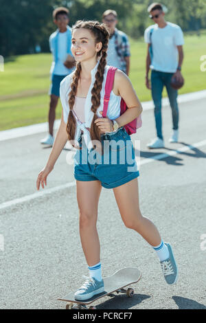 Smiling teenage Mädchen mit Rucksack, Skateboard, während Mitschüler hinter Walking im Park Stockfoto