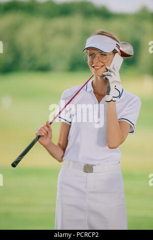 Portrait von lächelnd weibliche golf Spieler in Polo und Kappe mit Golf Club in der Hand sprechen auf dem Smartphone am Golfplatz Stockfoto