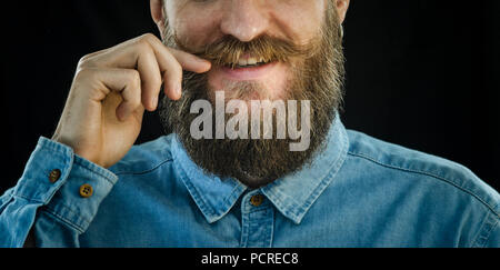 Lächelnd bärtigen Mann in ein blaues Jeanshemd Wendungen der Schnurrbart mit seiner Hand auf einem schwarzen Hintergrund. Hipster Look: Bart und Schnurrbart Close-up Stockfoto