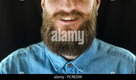Portrait von lächelnden bärtigen Mann in ein blaues Jeanshemd auf schwarzem Hintergrund. Hipster Look: Bart und Schnurrbart Close-up Stockfoto