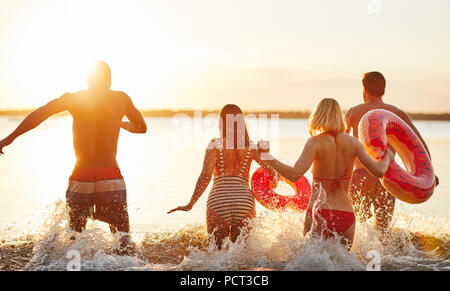 Rearview eine vielfältige Gruppe von jungen Freunden in Badebekleidung läuft in einen See und Spritzwasser zusammen bei Sonnenuntergang Stockfoto