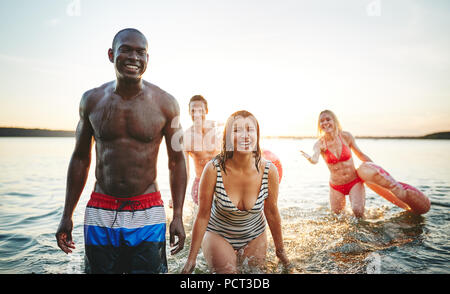 Gruppe von jungen Freunden in Badebekleidung Lachen und das Spritzen mit Wasser, während Spaß in einem See bei Sonnenuntergang Stockfoto