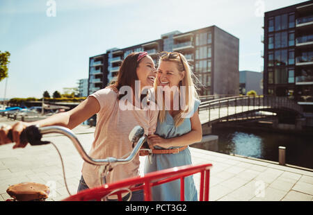 Zwei lachende Junge weibliche Freunde Spaß zu Fuß mit dem Fahrrad durch die Stadt im Sommer Stockfoto