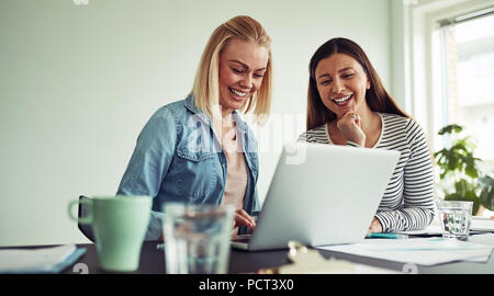 Zwei lächelnde junge Unternehmerinnen am Schreibtisch in einem Büro zusammen und reden mit einem Laptop Stockfoto