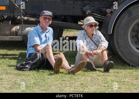Eine ältere oder ältere Paare in Shorts und Sonnenbrille schützen vor der Sonne und Schatten von einem geparkten Oldtimer in der heißen Sonne. Stockfoto