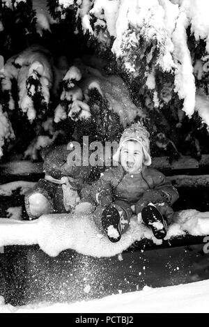 Kleiner Junge mit Teddy sitzt auf einer Bank im Schnee und Lachen Stockfoto