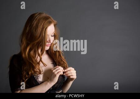 Eine rothaarige Mädchen steht im Studio trägt einen schwarzen lacy Kap. Sie sieht auf Ihr Haar in eine schüchterne Art und Weise. Stockfoto