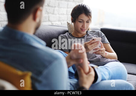 Junge hispanic Paar Festlegung auf der Couch zu Hause, mit einem Handy für Internet und Social Media. Der Freund ist der Massage nicht zu Füßen des Stockfoto