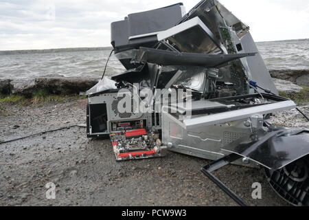 Defekten Computer, Monitore und Drucker vor der Natur Landschaft mit Fluss Hintergrund. Elektronische Geräte frei stehend in der Natur entsorgt. Computer Teile. Defekter Computer auf dem Boden im Freien Stockfoto