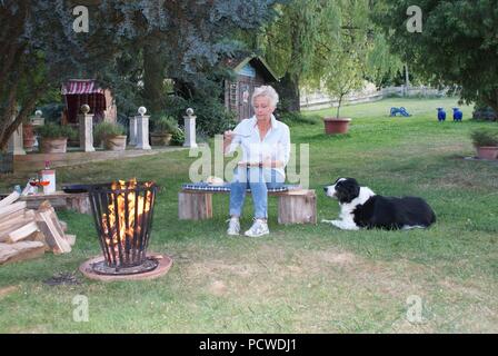 Die attraktive Frau isst Eier am Lagerfeuer gebraten, ihr Hund ist beobachten Stockfoto