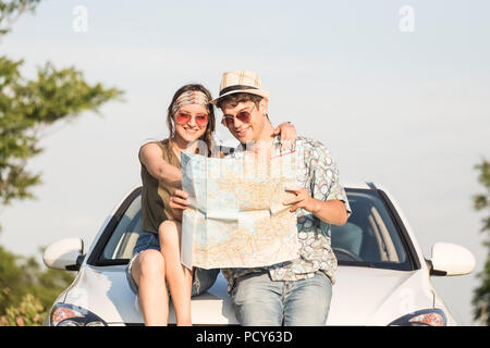 Junge hipster Paar sitzen auf Auto Motorhaube und Suchen auf der Karte. Fahrt auf der Straße. Stockfoto