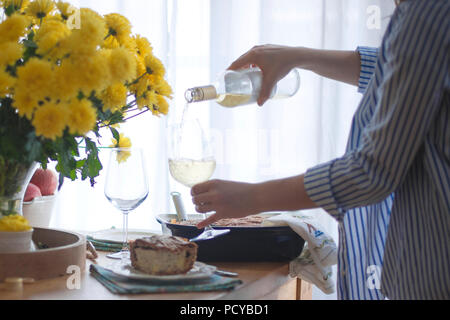 Abendessen für die Familie. Frau, die Portionen von Essen und Wein. Serviert Tisch. Gelbe Blumen auf dem Tisch. Tisch in der Nähe des Fensters. Stockfoto