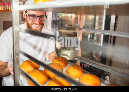 Ein junger hübscher baker Holding frische Brötchen mit Mohn im Hintergrund des Ofens. Stockfoto