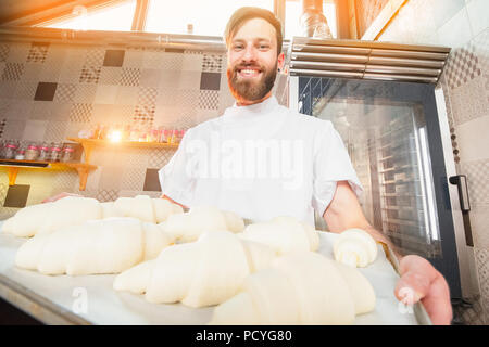 Ein junger hübscher Bäcker hält rohe Croissants von weißen Teig in den Händen eines Ofens. Halbzeuge aus Teig. Stockfoto