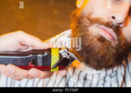 Friseur schneidet ein Bart mit einem Langhaarschneider auf ein junger hübscher Kerl mit einem Schnurrbart. Haarschnitt von einem Bart in einer männlichen Friseur. Stockfoto