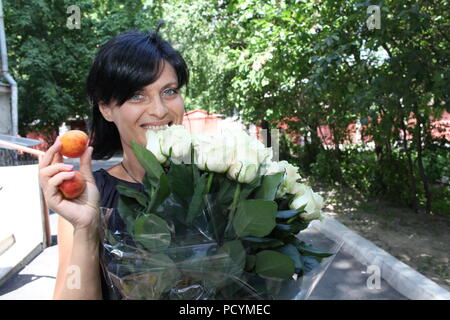 Junge hübsche Dame mit grauen Augen und einem großen Blumenstrauß aus den weißen Rosen und apriсots in der rechten Hand an einem schönen Sommermorgen am grünen Platz in Kiew Stockfoto