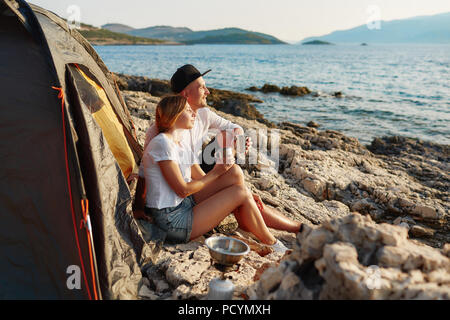 Seitenansicht des Romantisches Paar entspannend in der Nähe von Zelt bei Rock Beach, umarmen und bewundern Sie das Meer in den Sommerferien. Happy people Holding Bügeleisen Tasse Tee ich Stockfoto