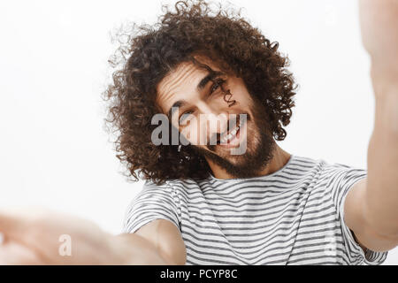 Close-up Portrait von positiven emotionalen hübscher Kerl mit Bart und stilvolle curly Haarschnitt, ziehen die Hände in Richtung Kamera und umarmen Sie, neigen, während ma Stockfoto