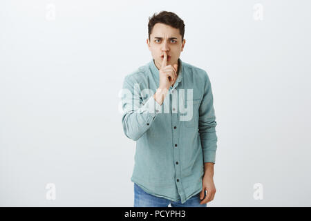 Ich bestelle Sie Stimme nach unten. Portrait von Missfallen ernsthaften jungen Mann in trendigen T-Shirt, sagen Shh, psst Geste mit dem Zeigefinger über Mund, Stockfoto