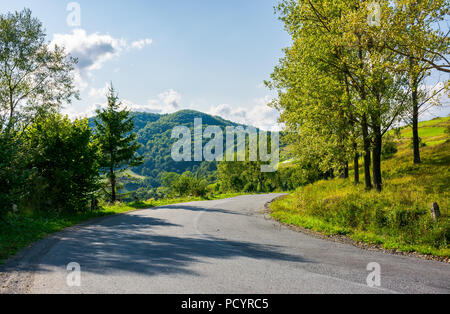 Land Straße den Hügel hinunter. Wald entlang des Weges und die Berge in der Ferne. Wunderbares sonniges Wetter im frühen Herbst Stockfoto